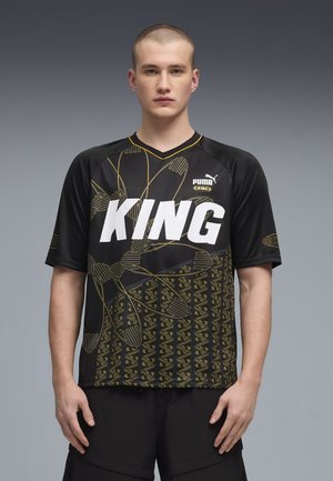 Junger Mann trägt schwarzes Puma King Sporttrikot mit goldenen geometrischen Mustern und klarem weißen "KING"-Schriftzug, steht vor grauem Hintergrund.