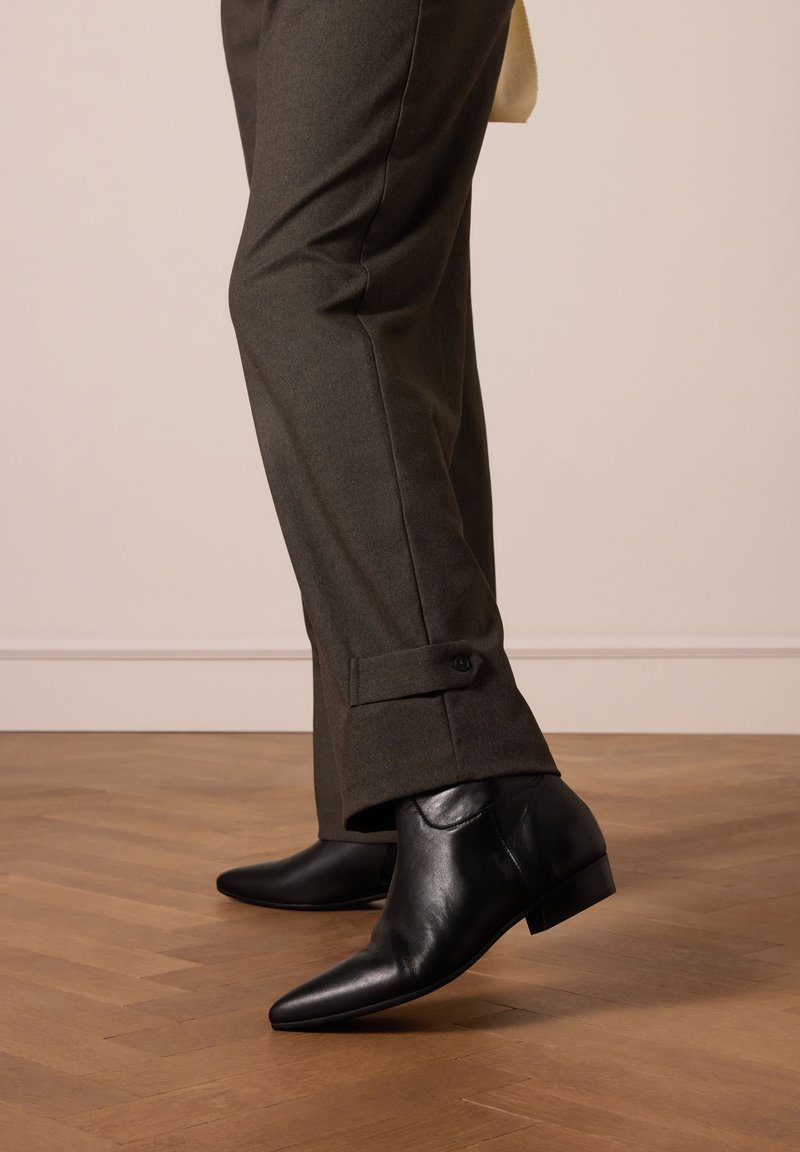 Schwarze Leder-Stiefeletten mit spitzer Zehenform und niedrigem Absatz, kombiniert mit dunkelgrünen Hosen, die über einen gepolsterten Knopfverschluss verfügen.