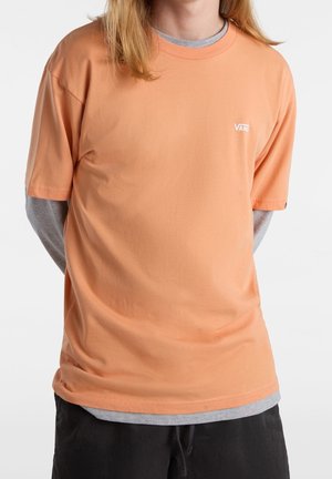 Pomarańczowy t-shirt z krótkim rękawem wykonany z bawełny, z okrągłym dekoltem i małym białym logo „VANS” na lewej piersi. Nałożony na szarą koszulkę pod spodem.