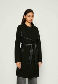 Cappotto nero con colletto drappeggiato, cintura di pelle e tasche frontali. Realizzato in un tessuto morbido, presenta un design avvolgente contemporaneo.
