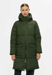Vrouw in een lange, donkergroene gewatteerde winterjas met een capuchon, een rits aan de voorkant en twee grote zakken aan de voorkant, staande voor een effen achtergrond.