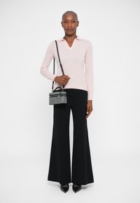 Maglietta a maniche lunghe rosa chiaro con scollatura a V e collo, abbinata a pantaloni neri a gamba larga. La modella tiene in mano una piccola borsa nera con tracolla.