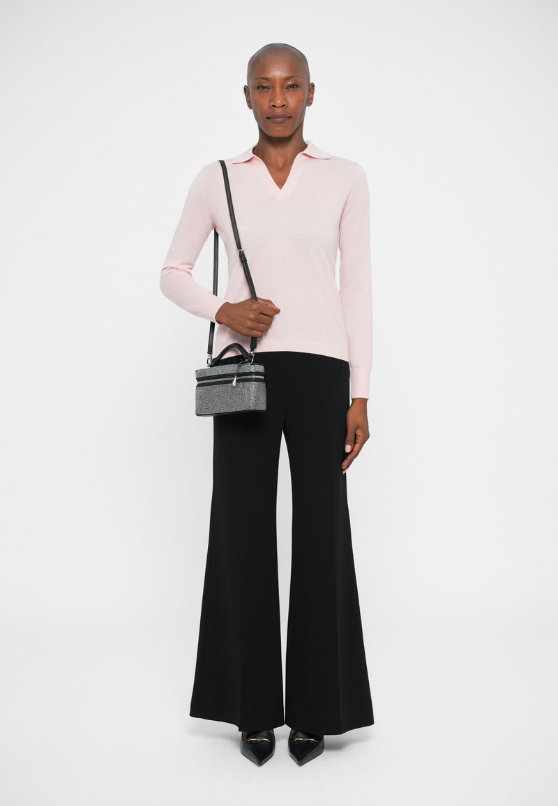 Maglietta a maniche lunghe rosa chiaro con scollatura a V e collo, abbinata a pantaloni neri a gamba larga. La modella tiene in mano una piccola borsa nera con tracolla.