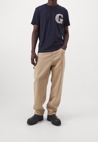 Camiseta de algodón azul marino con un gran gráfico de "G", combinada con pantalones sueltos de color beige claro y zapatos de tacón grueso negros.