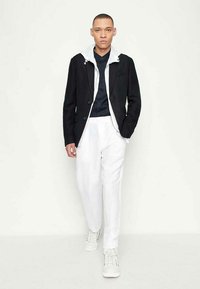 Blazer nero sopra una camicia blu navy, abbinato a pantaloni bianchi e sneakers bianche. Il blazer ha un cappuccio grigio e accessori in metallo argentato.