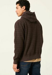 Brauner Hoodie mit strukturiertem Muster, ausgestattet mit einer Kordelzugkapuze und gerippten Bündchen. Kombiniert mit dunkelblauen Jeans.