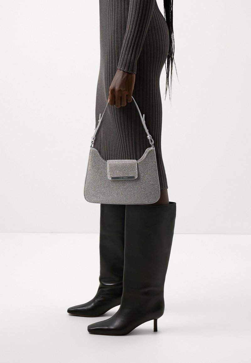 Femme en robe côtelée foncée et bottes noires à talons montant aux genoux, tenant un petit sac à main argenté sur un fond clair uni.