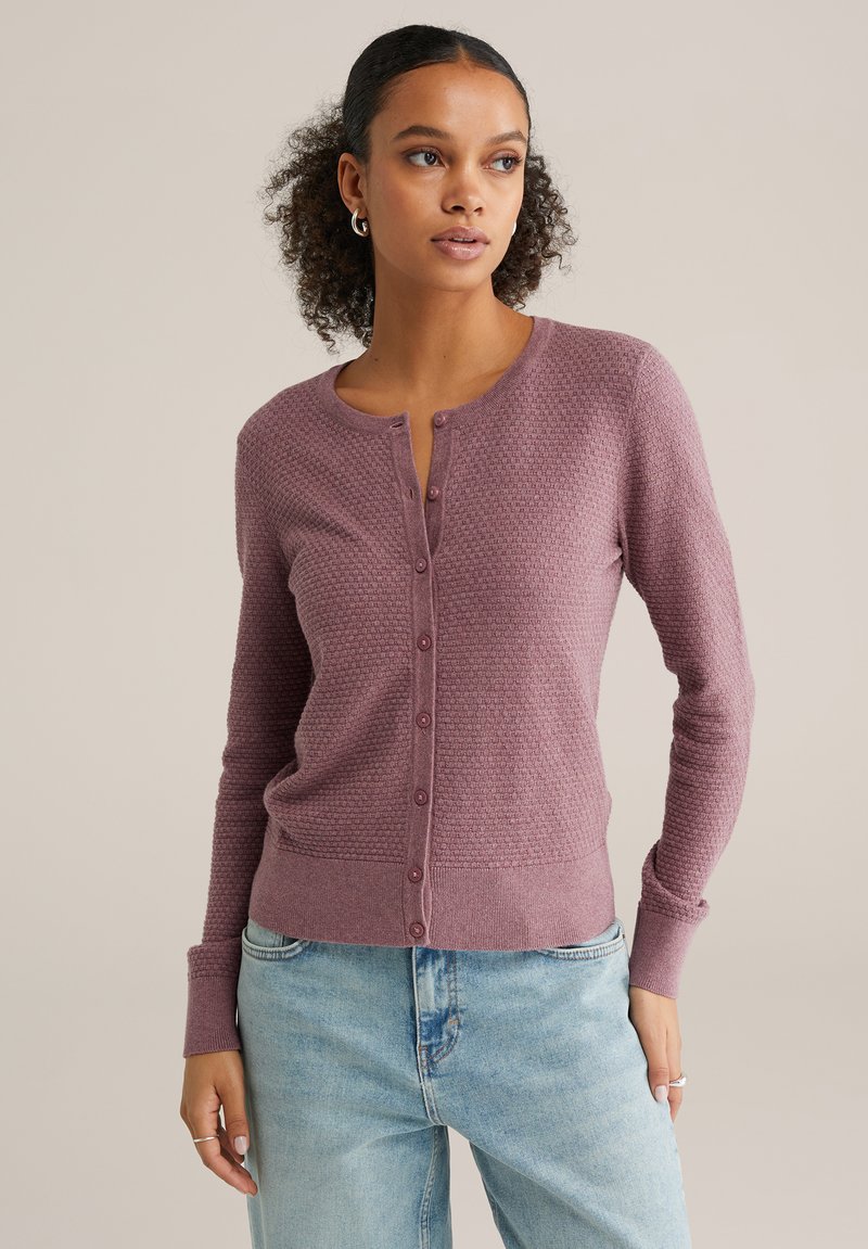 Langermige, gestructureerde cardigan in gedempte mauve met een knoopsluiting aan de voorkant en geribbelde manchetten. Gecombineerd met lichtblauwe jeans.