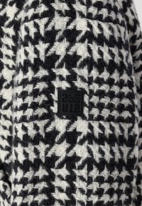 Ein schwarz-weißer Hahnentrittmuster-Stoff mit strukturierter Oberfläche und einem kleinen schwarzen Logopatch an der Seite.