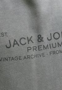 Tissu gris avec un texte imprimé en noir indiquant « JACK & JONES PREMIUM VINTAGE ARCHIVE » en disposition courbée.