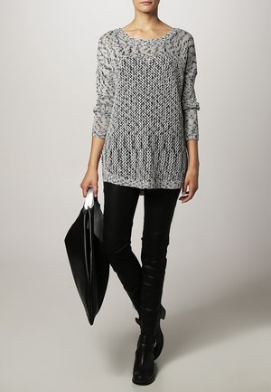 Vrouw draagt een grijze gebreide trui, zwarte leren broek, zwarte laarzen en houdt een grote zwarte leren totebag vast.