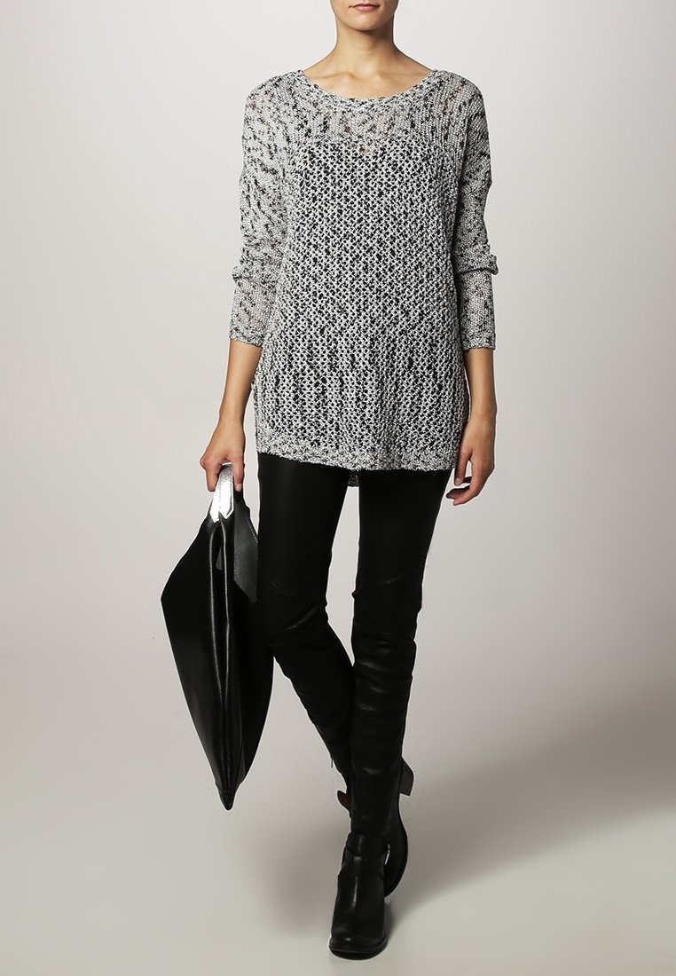 Femme portant un pull en maille gris, un pantalon en cuir noir, des bottes noires, tenant un grand sac fourre-tout en cuir noir.