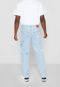 Pantaloni cargo in denim azzurro chiaro con molteplici tasche, una toppa in pelle sul retro e dettagli del logo bianchi, abbinati a una t-shirt bianca.