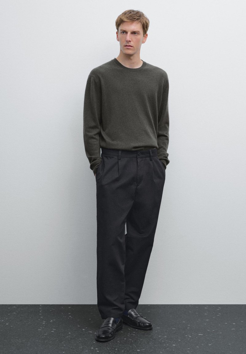 Tmavě zelený pletený svetr, volného střihu, zkombinovaný s černými kalhotami s vysokým pasem. Černé mokasíny doplňují outfit na neutrálním pozadí.