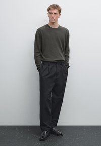 Dunkelgrüner, langärmeliger Pullover aus einem weichen Stoff, kombiniert mit schwarzen, weit geschnittenen Hosen und schwarzen Slippern. Neutrale Hintergrund.