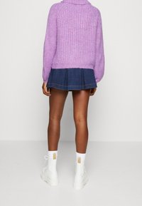 Personne portant un pull violet en tricot, une jupe plissée courte bleu foncé, des chaussettes blanches, et des bottes blanches à lacets, debout sur un fond uni.
