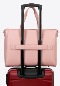 Bolso tote sintético rosa con asas superiores descansando sobre una maleta dura roja, asegurada por un mango de carro negro. Superficie texturizada visible.