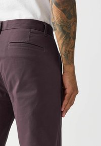 Dunkelviolette Chinos mit glatter Textur, ausgestattet mit einer Gesäßtasche und schlankem Schnitt, vor einem neutralen Hintergrund präsentiert.