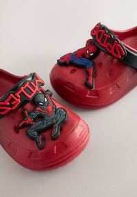 Zuecos rojos con correas negras que presentan figuras en relieve de Spiderman y Spider-Man. Hechos de caucho, tienen agujeros de ventilación y suelas texturizadas.