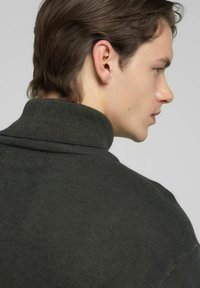 Suéter de cuello alto verde oscuro con textura acanalada. Presenta un cuello alto y detalles sutiles de costura, mostrado desde atrás.