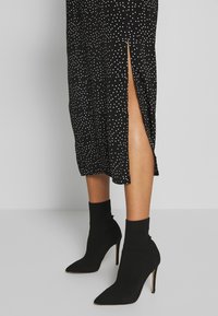 Vestido midi negro con lunares blancos, con abertura lateral, combinado con botas de tobillo negras de tacón stiletto y punta afilada, con diseño de calcetín acanalado.