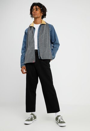 Chaqueta de mezclilla y forro polar gris con cuello beige, camiseta blanca debajo, pantalones de pierna ancha negros y zapatillas de ante verdes.