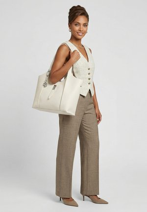 Donna con gilet beige e pantaloni marroni che porta una grande borsa bianca, sorridente, in piedi davanti a uno sfondo chiaro e semplice.