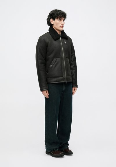 Juoda odinė striukė su kailiniu apykakle, užtrauktuko užsegimu ir dviem šoninėmis kišenėmis; derinama su tamsiai mėlynomis džinsais ir rudu batais.