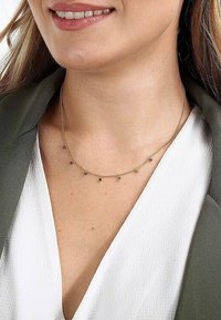 Goldene Kettenhalskette mit kleinen, mehrfarbigen Edelsteinakzenten, die in einem zarten Muster angeordnet sind, getragen über einer weißen Bluse und einem grünen Blazer.