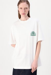 White cotton t-shirt with short sleeves, featuring a green graphic print on the left chest area. Classic crew neck design.