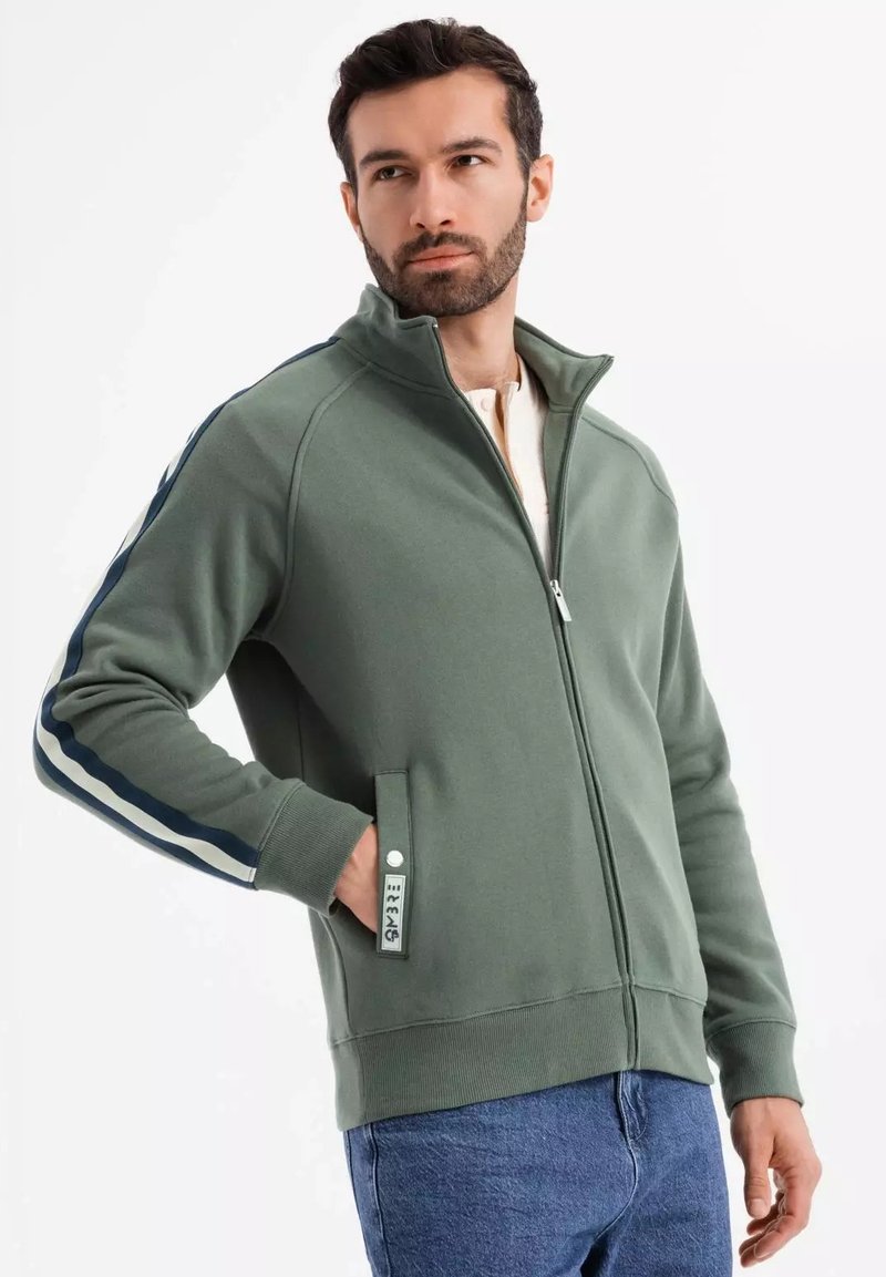 Veste zippée verte en tissu doux, avec un col haut, des poches latérales et des accents rayés bleu marine sur les manches.