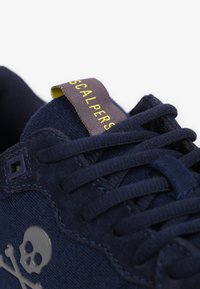 Marineblauwe sneaker met een bovenwerk van suede en stof, met een gestructureerd oppervlak, doodskopembleem en een lipje met gele accenten.