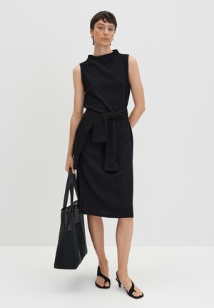 Robe noire sans manches avec une taille cintrée par un lien, tissu texturé, silhouette mi-longue, associée à un sac en maille et des sandales.