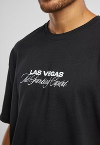 Sort t-shirt med hvid broderet tekst, der står "LAS VEGAS The Gambling Capital." Enkelt crew neck design med korte ærmer.