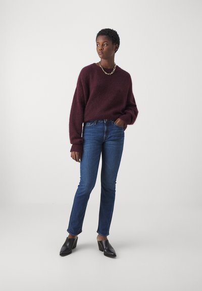 Suéter de punto burdeos de gran tamaño con puños acanalados; jeans azules de corte ajustado; mules negros de tacón. El modelo posa con una mano en el bolsillo.
