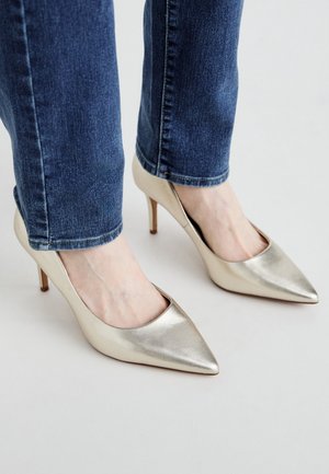 Pieds portant des escarpins à bout pointu en métal doré associés à un jean en denim bleu sur un fond blanc uni.