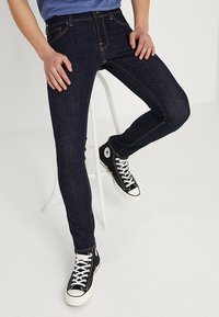 Mörkblå slim-fit jeans med orange sömnadsdetaljer, tillsammans med svarta högklackade sneakers med vita snören och en cirkulär logotyp.