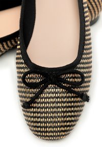 Close-up van een zwart en beige geweven ballerina met zwarte rand en strik op de teen tegen een witte achtergrond.