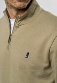 Beige zip-up hettegenser laget av mykt stoff, med høy krage og en liten marineblå brodert logo på venstre bryst.