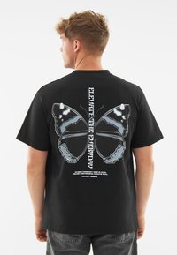 Černé bavlněné tričko s grafickým motivem motýla na zádech, bílým textem a kontrastními modrými detaily, krátkými rukávy a volným střihem.