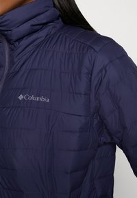 Granatowa, ocieplana kurtka z poziomymi przeszyciami, wysokim kołnierzem i logo Columbia na piersi. Gładki, lekki materiał o nowoczesnym designie.
