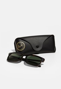 Ochelari de soare Ray-Ban cu lentile verzi închise într-un cadru din tortoiseshell, poziționați lângă un etui de protecție texturat, de culoare neagră, cu detalii ale logo-ului.