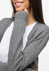 Gray knit cardigan with ribbed cuffs, featuring a lightweight, soft texture. Worn over a white top, showing a relaxed fit and open design.