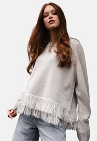 Junge Frau mit langen braunen Haaren, die einen hellgrauen Pullover mit Fransen am Saum und hellblauen Jeans trägt, und vor einem einfarbigen Hintergrund steht.