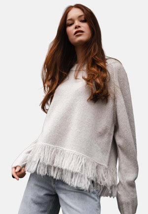 Junge Frau mit langen braunen Haaren, die einen hellgrauen Pullover mit Fransen am Saum und hellblauen Jeans trägt, und vor einem einfarbigen Hintergrund steht.