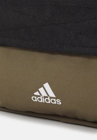 Svart quiltad tyg möter olivgrön texturerad material, med en vit Adidas-logotyp på den nedre delen. Kompakt och hållbar design.
