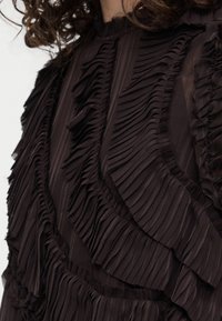 Κοντινή λήψη σκουρόχρωμης μπλούζας με στιβαρές κάθετες βολάν και πιέτες στο στήθος και τα μανίκια. Μερικώς ορατά κυματιστά μαλλιά.