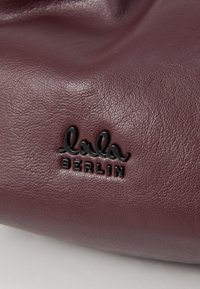 Bordo odinės rankinės, su tekstūruotu paviršiumi ir ryškiu juodu logotipu, kuriame užrašyta "lala BERLIN", išspaudytu medžiagoje.