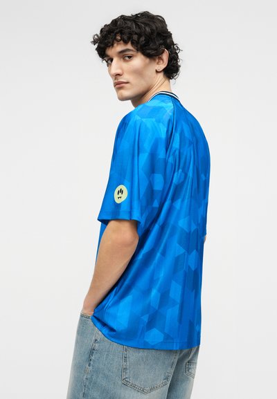 Νεαρός με κοντά σγουρά μαλλιά που φοράει μπλε πουκάμισο με γεωμετρικό σχέδιο και ανοιχτόχρωμο τζιν, κοιτάζει πάνω από τον ώμο προς τα αριστερά.