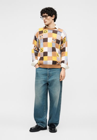 Νέος άνδρας με σγουρά μαλλιά που φοράει γυαλιά, πουλόβερ με καρό σχέδιο, αρκουδάκια και προσωπάκια, μπλε τζιν και μαύρα παπούτσια σε λευκό φόντο.